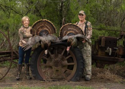 A woman and a man in camouflage clothing stand next to an old tractor wheel, each holding a wild turkey. The turkeys hang over the wheel with their feathers fanned out. Surrounding them is lush greenery.