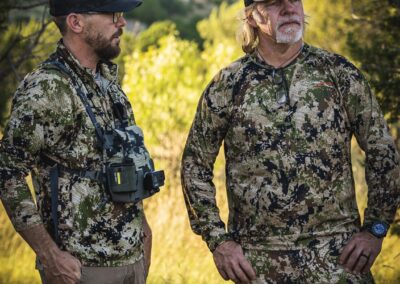 Two individuals dressed in camouflage clothing are positioned in a field, emphasizing their connection to the outdoor environment.