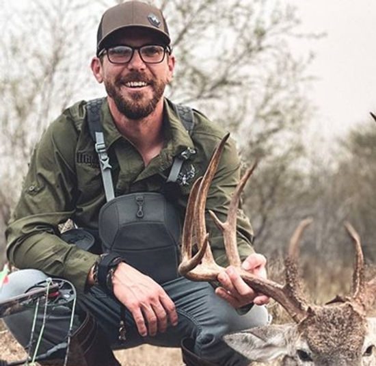 A man wearing glasses, a cap, and outdoor gear is kneeling and smiling while holding the antlers of a deer he has hunted. He is surrounded by dry vegetation and leafless trees in the background.