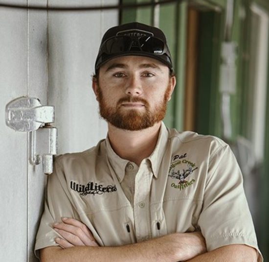 A man with a beard and mustache stands with his arms crossed, wearing a beige shirt with various embroidered logos and a cap with sunglasses perched on top. In the background, there is a green wall with a metallic latch.