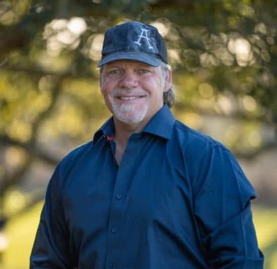 A smiling man wearing a black baseball cap and dark blue button-up shirt stands outdoors with trees in the background.