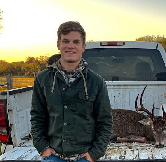 A man stands smiling in front of a white pickup truck parked in an outdoor setting. In the truck bed is a large buck deer with antlers. The man is wearing a dark green jacket and jeans, with his hands in his pockets. The background features a yellow and orange sunset.